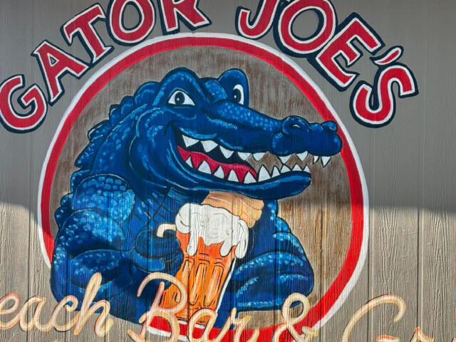 Sign for Gator Joe's with a blue alligator holding a beer inside a circular red-and-white logo, blue sky above.