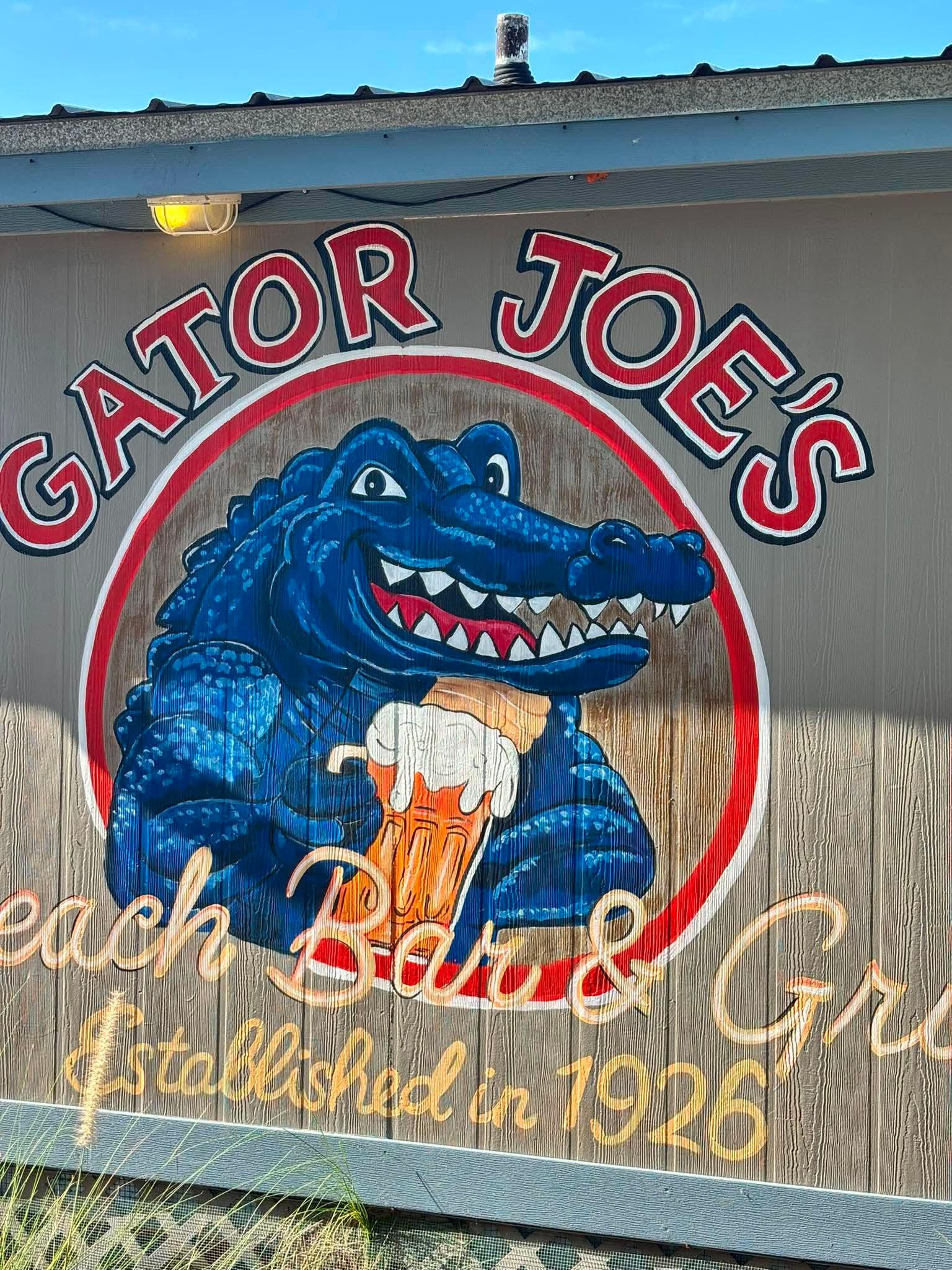 Sign for Gator Joe's with a blue alligator holding a beer inside a circular red-and-white logo, blue sky above.