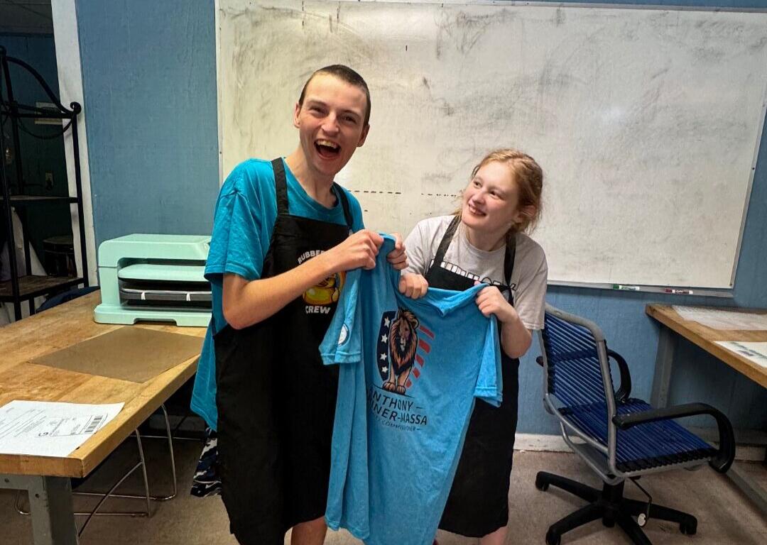 Two people in aprons smile while proudly holding up a blue T-shirt with a lion crest design in a room with a whiteboard behind them.