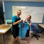 Two people in aprons smile while proudly holding up a blue T-shirt with a lion crest design in a room with a whiteboard behind them.