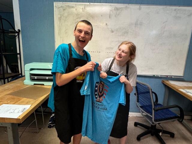 Two people in aprons smile while proudly holding up a blue T-shirt with a lion crest design in a room with a whiteboard behind them.