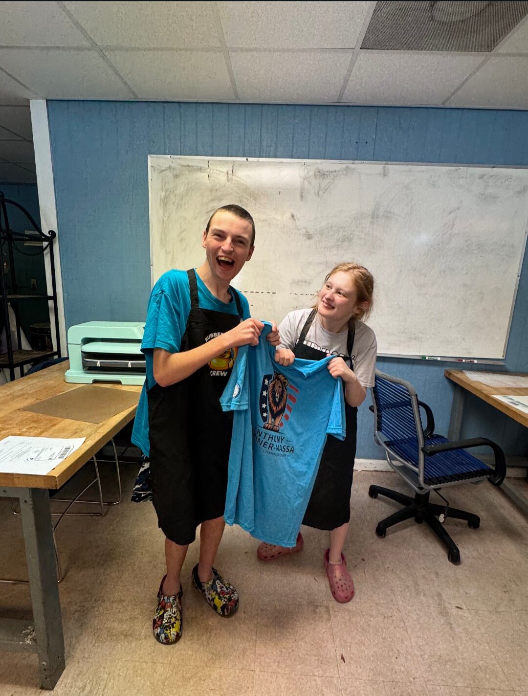 Two people in aprons smile while proudly holding up a blue T-shirt with a lion crest design in a room with a whiteboard behind them.