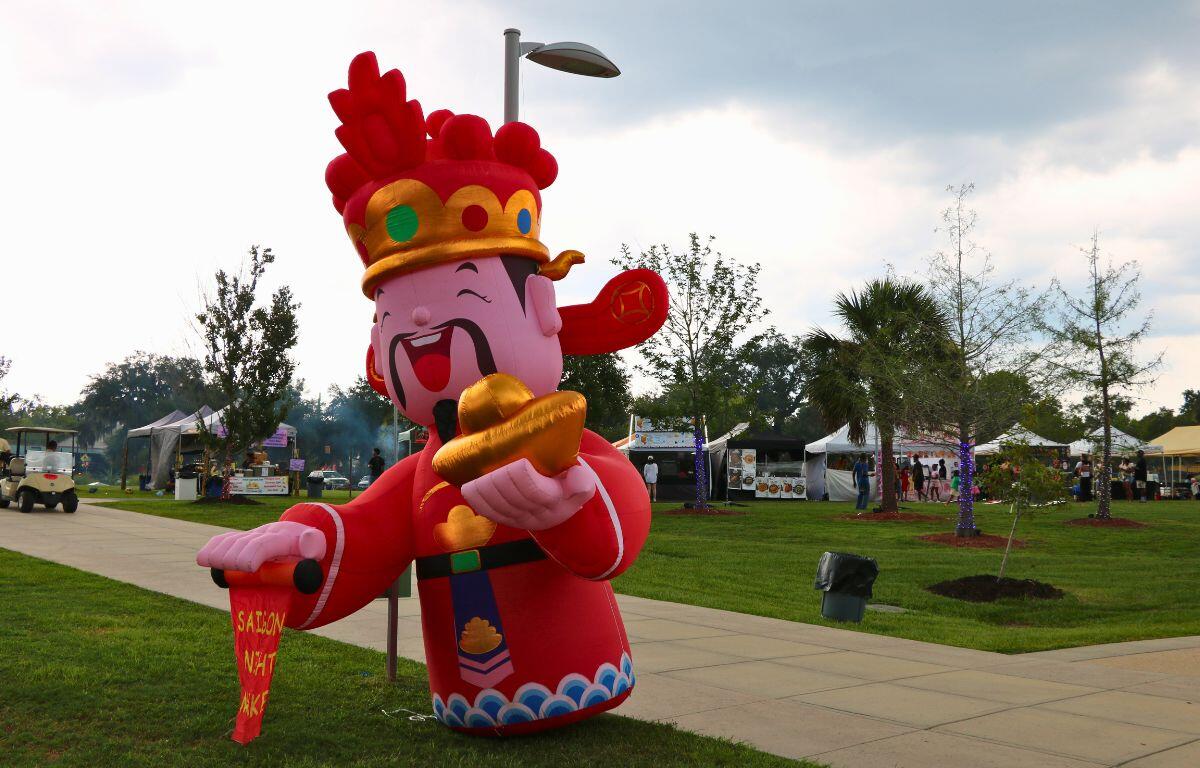 Inflatable cartoon Chinese deity in red robes holding gold ingots at an outdoor festival park with tents in the background.