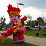 Inflatable cartoon Chinese deity in red robes holding gold ingots at an outdoor festival park with tents in the background.