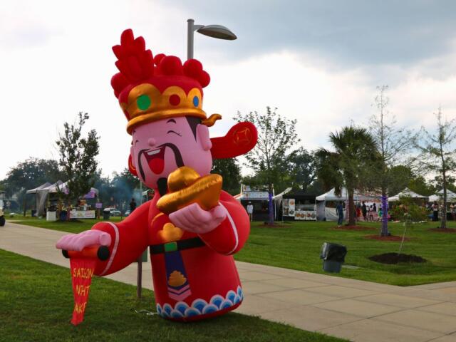 Inflatable cartoon Chinese deity in red robes holding gold ingots at an outdoor festival park with tents in the background.