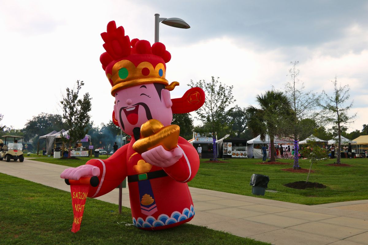 Inflatable cartoon Chinese deity in red robes holding gold ingots at an outdoor festival park with tents in the background.