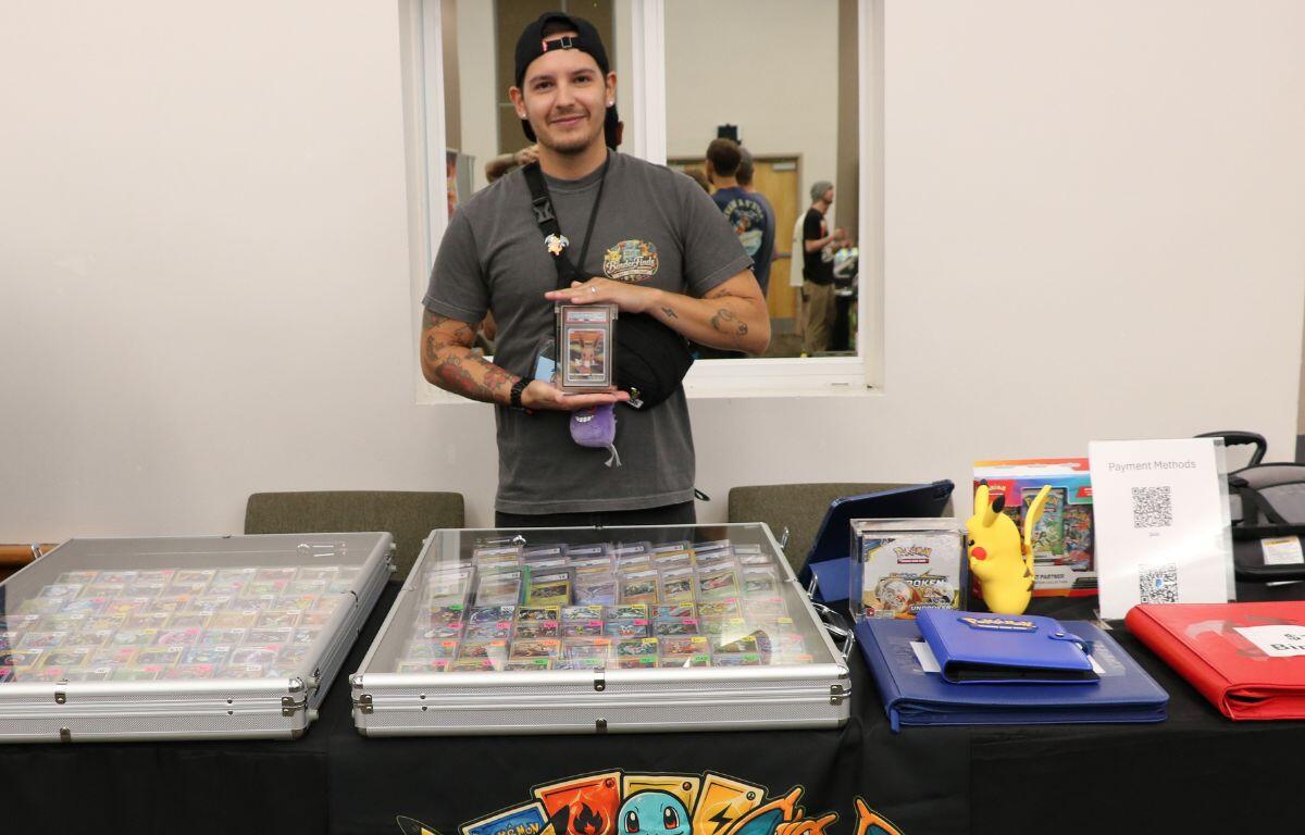 Vendor in a gray T-shirt and backward cap presents trading cards in clear cases at a Pokémon card booth.