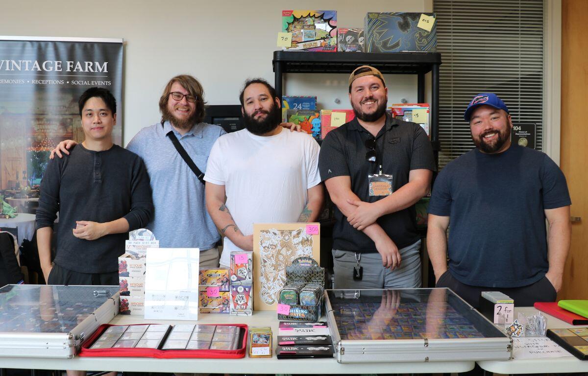 Five men standing behind a table of games and trading cards at a booth with a Vintage Farm banner visible on the left.