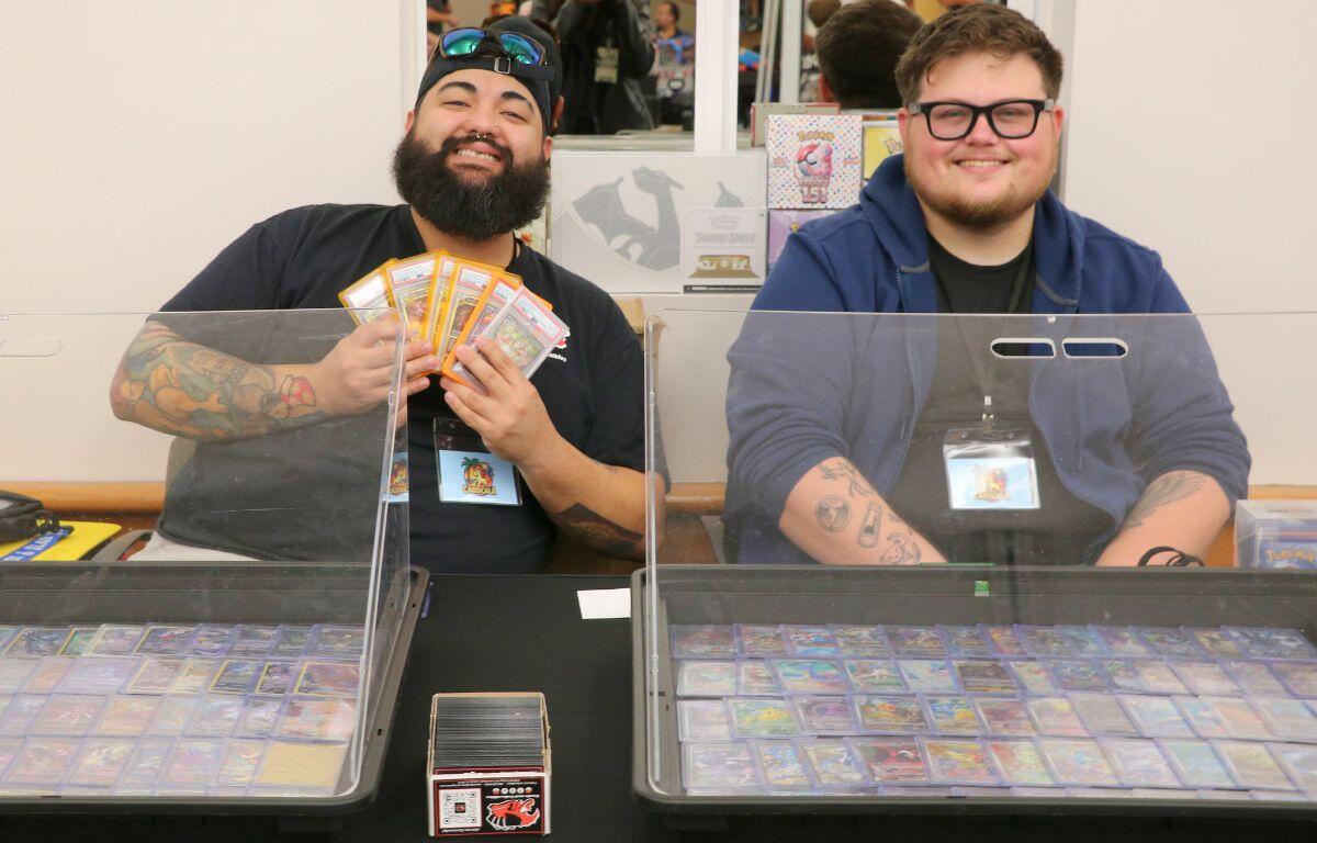 Two men sit together, smiling. The man on the left holds multiple trading cards.