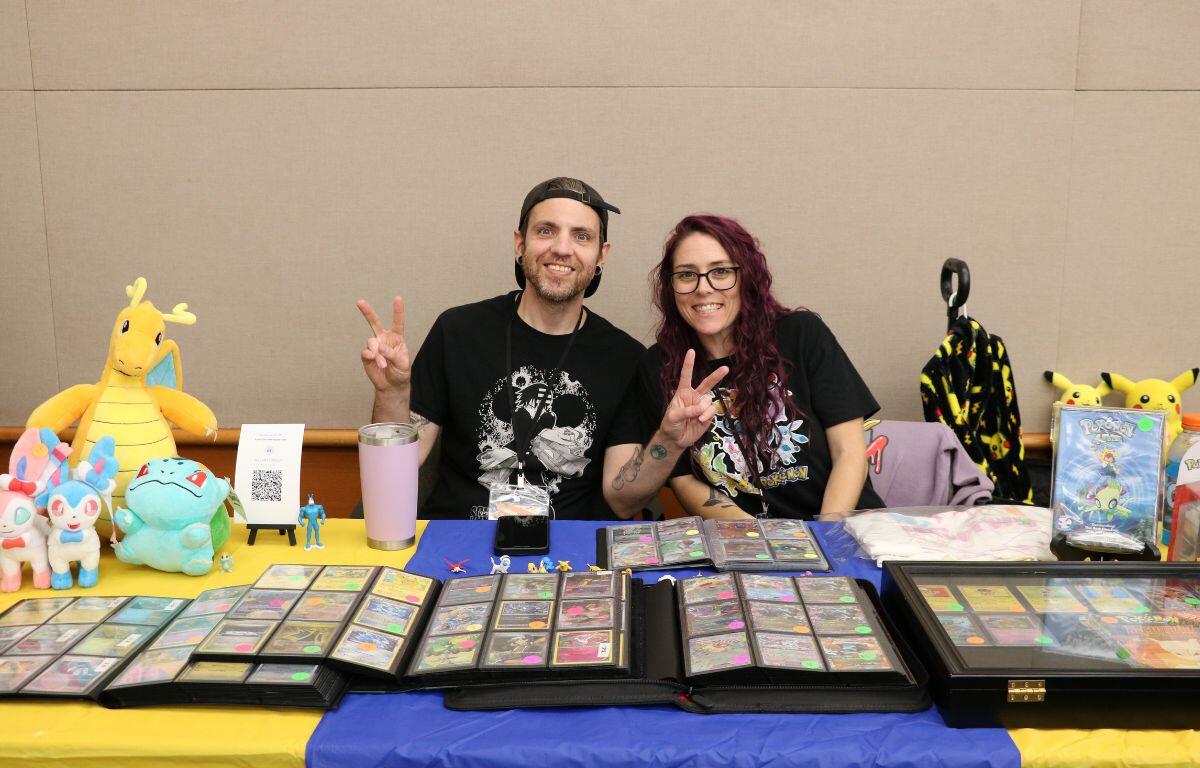 Two smiling vendors sit behind a table of colorful trading cards in display binders, with plush toys and Pokemon merchandise nearby.