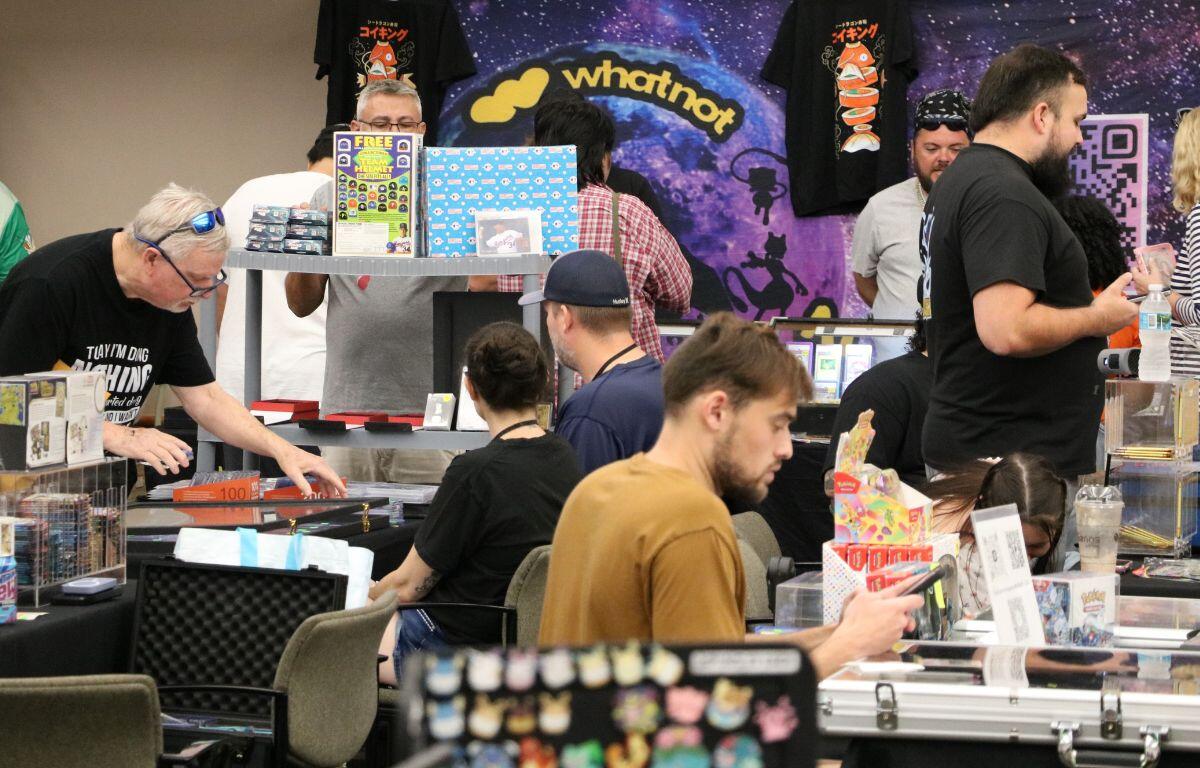Vendor at a convention booth displaying trading cards, with a WhatNot backdrop and shoppers at tables.