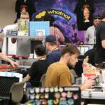 Vendor at a convention booth displaying trading cards, with a WhatNot backdrop and shoppers at tables.