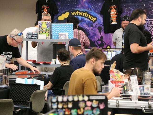 Vendor at a convention booth displaying trading cards, with a WhatNot backdrop and shoppers at tables.