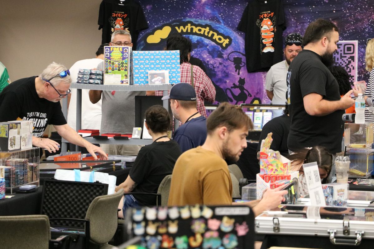 Vendor at a convention booth displaying trading cards, with a WhatNot backdrop and shoppers at tables.
