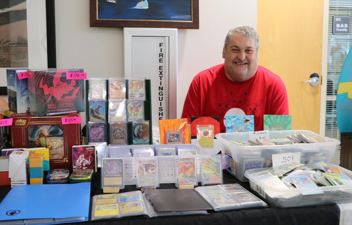 Smiling man in a red shirt sits behind a table full of Pokémon cards and display boxes for sale.