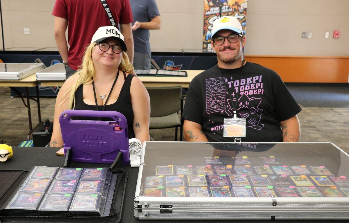 Two people sit at a table with trading cards displayed in open cases, wearing hats and lanyards at a convention booth.