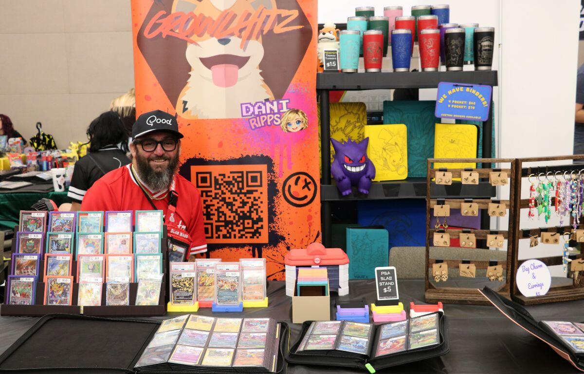 Vendor sits behind a table of trading cards at a convention with a bright orange banner of a cartoon dog and QR code.