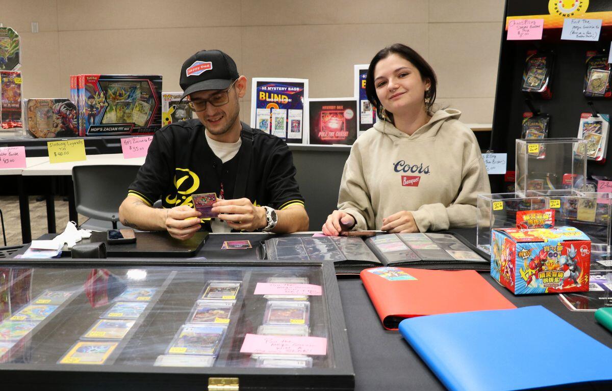 Two people sit at a table organizing trading cards with card cases and displays around them at a convention.