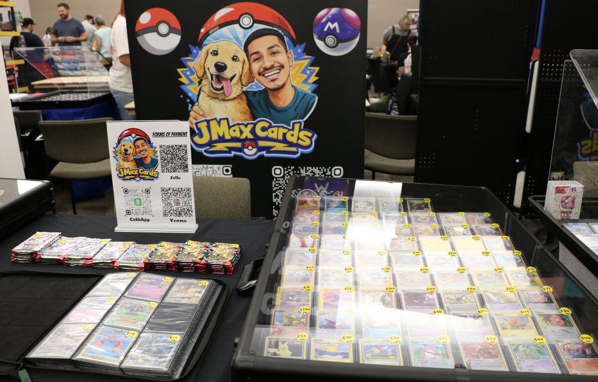 At a convention booth for JMaxCards, a man and dog cartoon mascot on a banner with pokéball icons behind a glass card display.