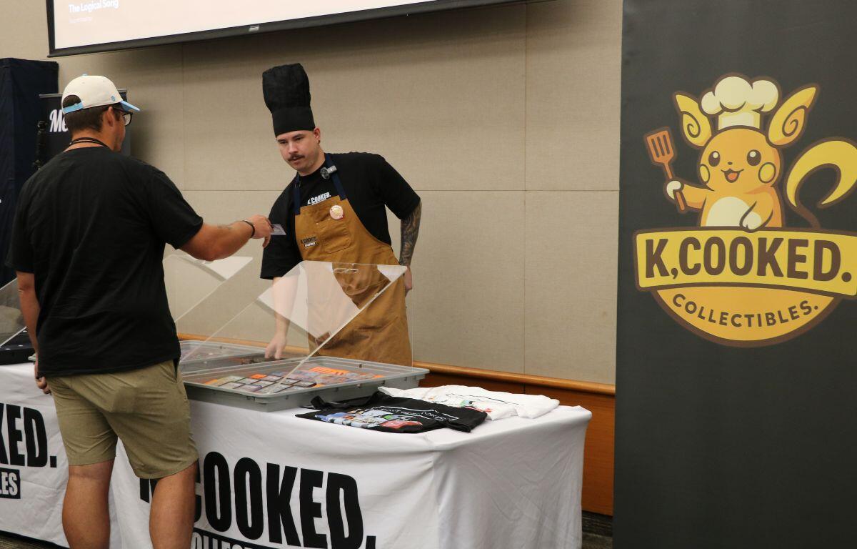 Customer wearing a white cap hands a card to a vendor in a tall chef’s hat at the K.Cooked Collectibles booth.