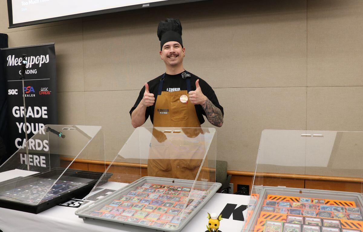 Trading card booth owner wears black chef's hat and a brown apron while showcasing trading cards in large, clear cases.