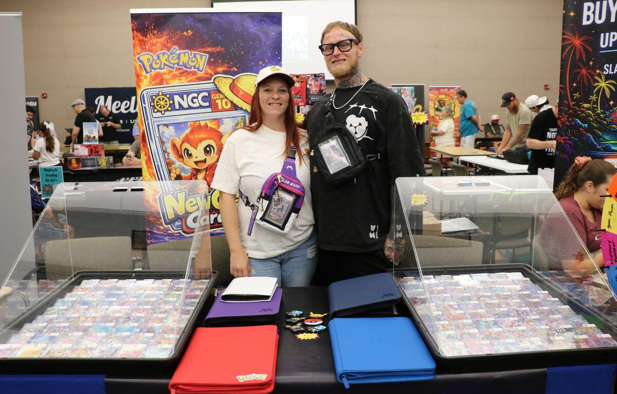 Two people smile at a Pokémon card booth, with glass display cases of cards and colorful binders on the table behind them.