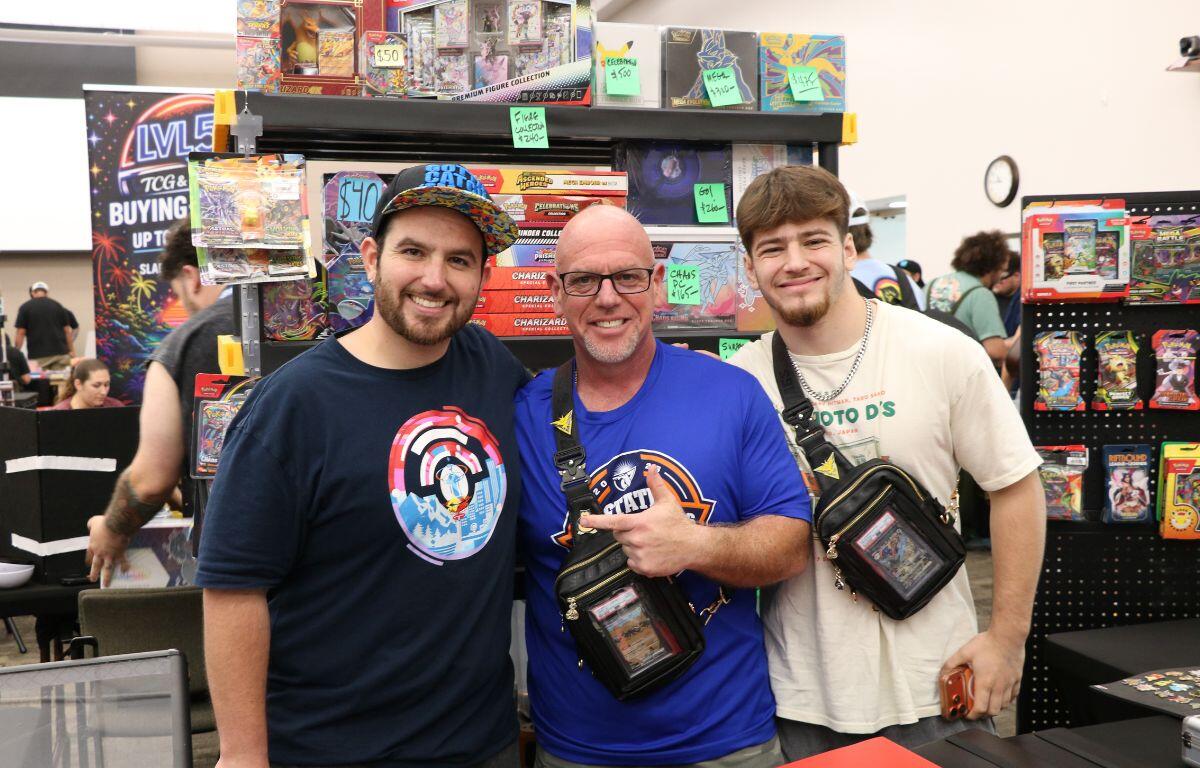 Three men posing together at a toy booth with shelves of boxed action figures behind them.