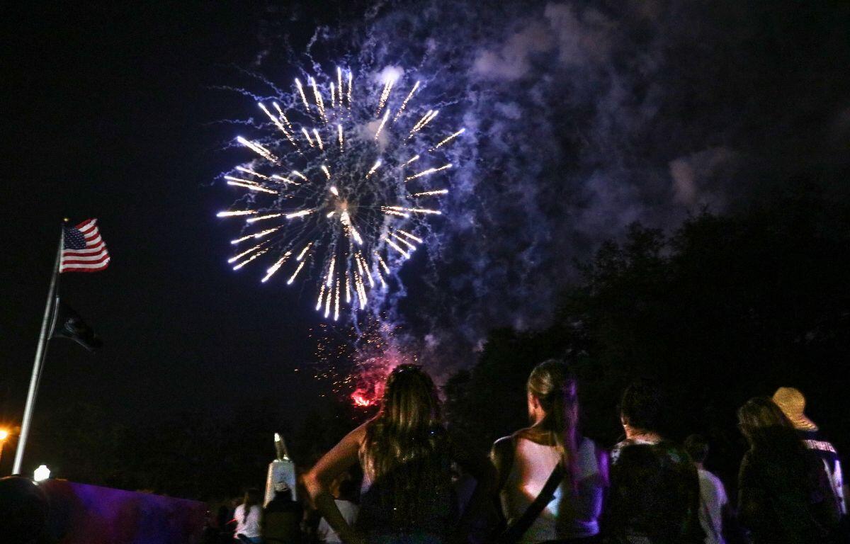 Fireworks explode in the night sky over a crowd of spectators, with an American flag on a pole to the left.