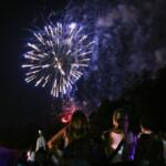 Fireworks explode in the night sky over a crowd of spectators, with an American flag on a pole to the left.