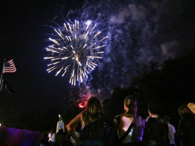 Fireworks explode in the night sky over a crowd of spectators, with an American flag on a pole to the left.