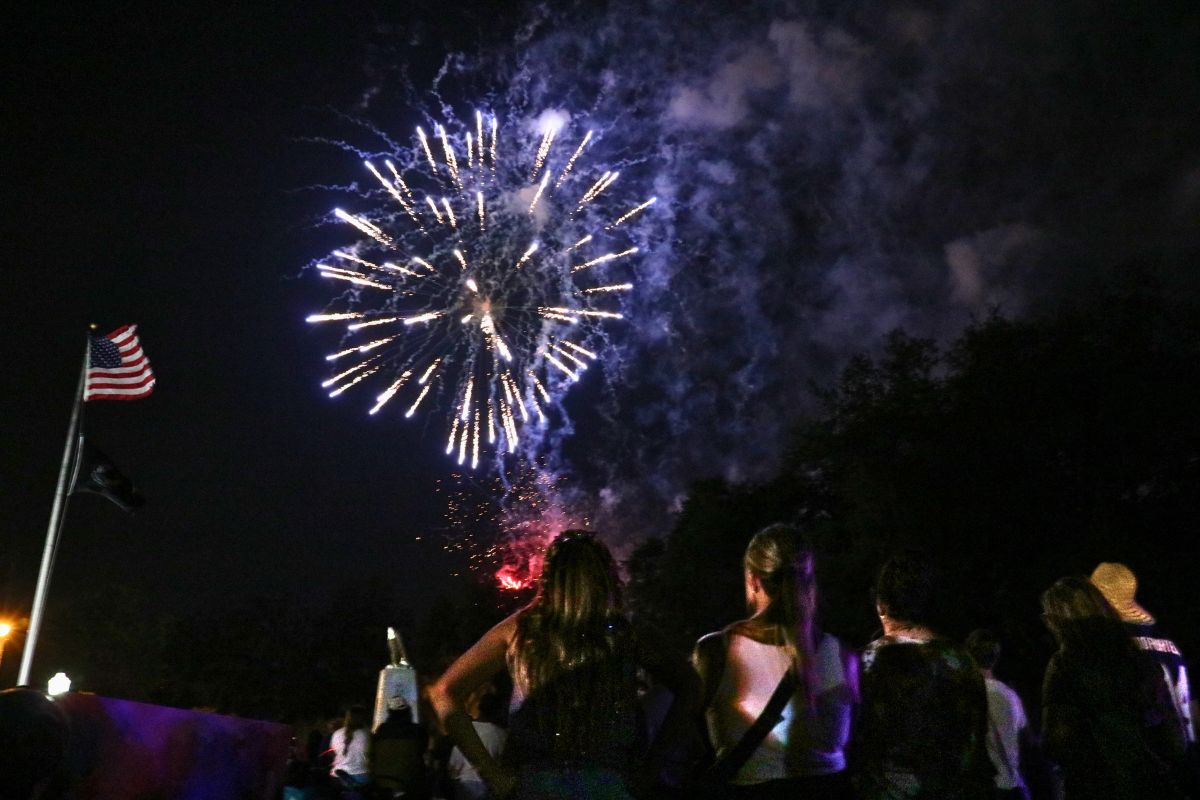 Fireworks explode in the night sky over a crowd of spectators, with an American flag on a pole to the left.