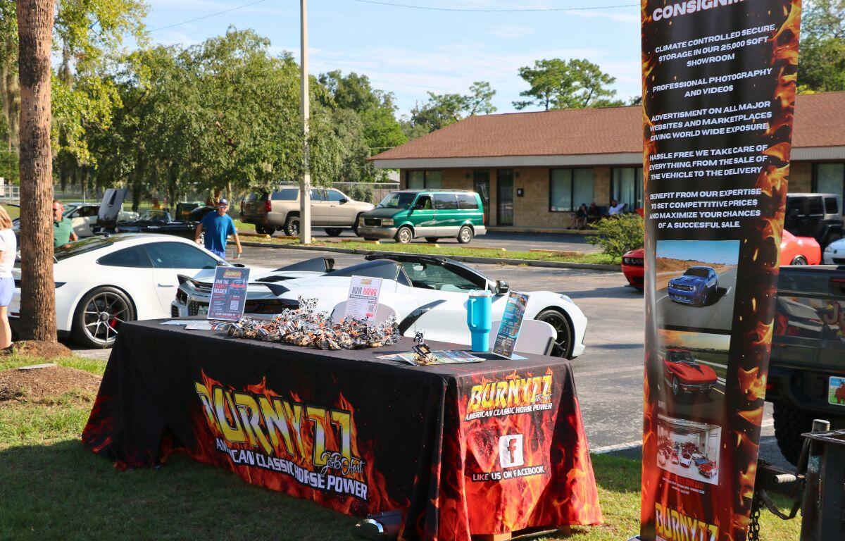 Outdoor car show display: a table with promotional materials and a tall flaming banner beside a white sports car and a green van in the background.