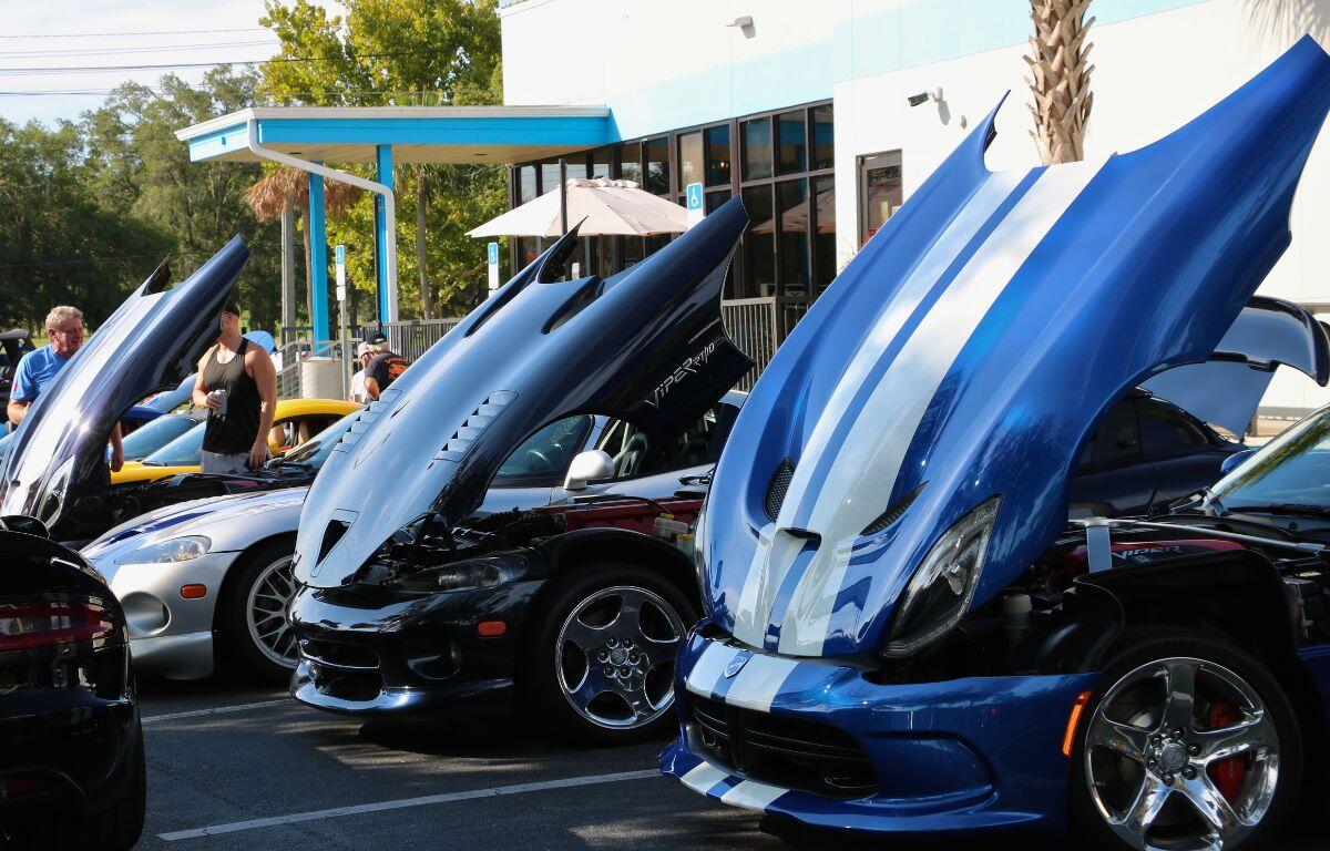 Row of blue and black sports cars with raised gull-wing doors at an outdoor car show, with people walking nearby.