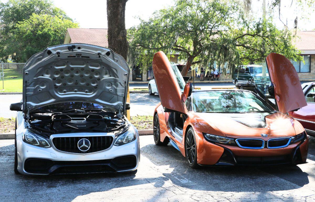 Two luxury cars at a car show: white Mercedes with hood open next to orange BMW i8 with gullwing doors raised.