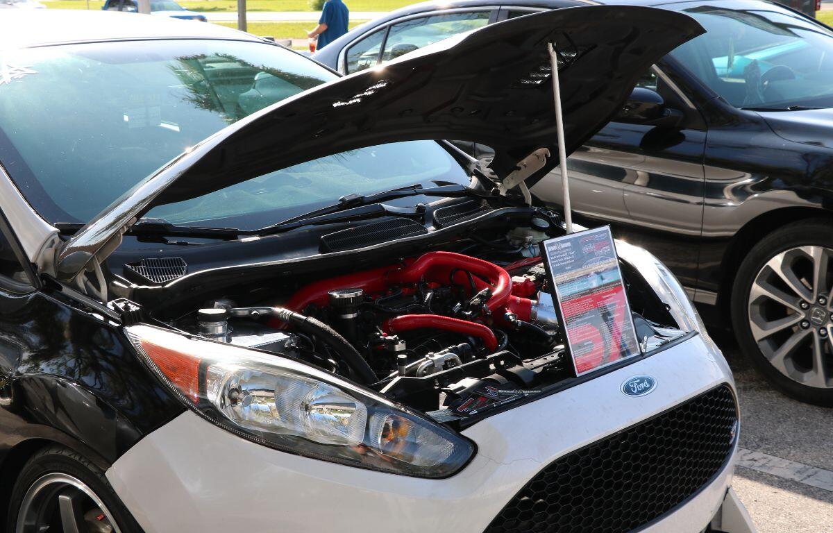 Hood up on a Ford during a car show, exposing a red aftermarket engine with an information sheet propped beside it.