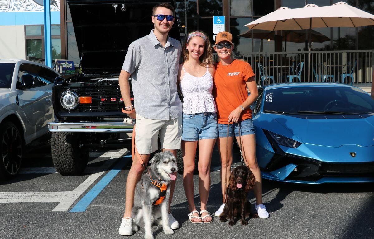 Three friends posing with two dogs in a parking lot next to a blue sports car and a black vintage jeep, smiling at the camera on a sunny day.