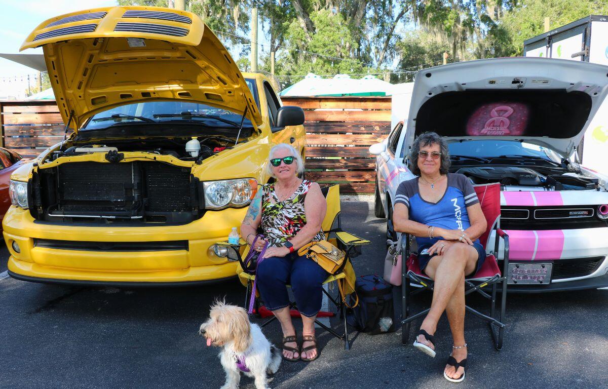 Two women sit in folding chairs between a yellow pickup with its hood raised and a white car with a pink stripe and hood open, a small dog at their feet.