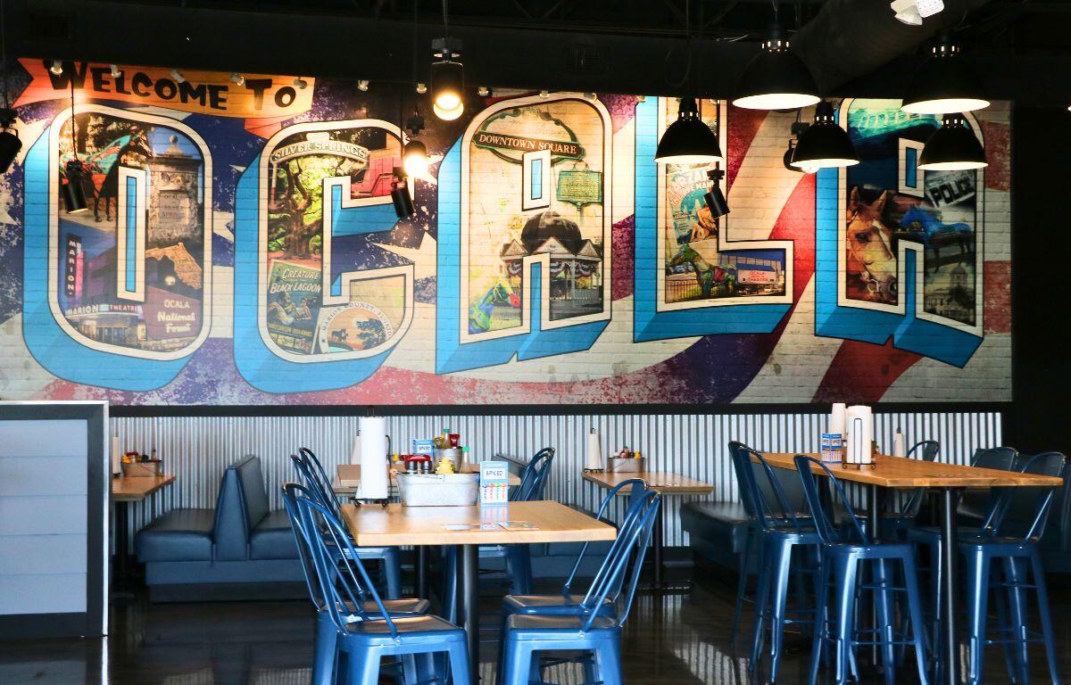 Colorful mural spelling 'OCALA' on a brick wall behind blue metal chairs and wooden tables in a casual restaurant.