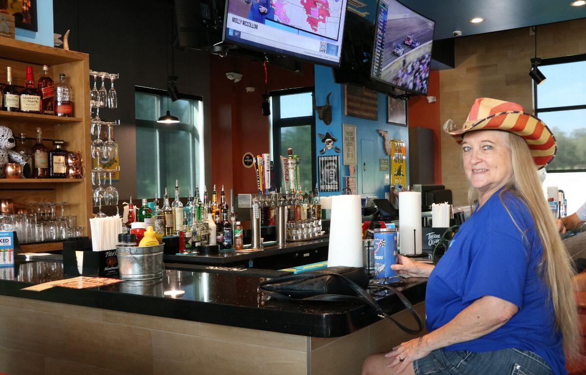 Smiling woman in a blue shirt and striped cowboy hat sits at a bar with bottles behind her on the shelves and taps nearby.