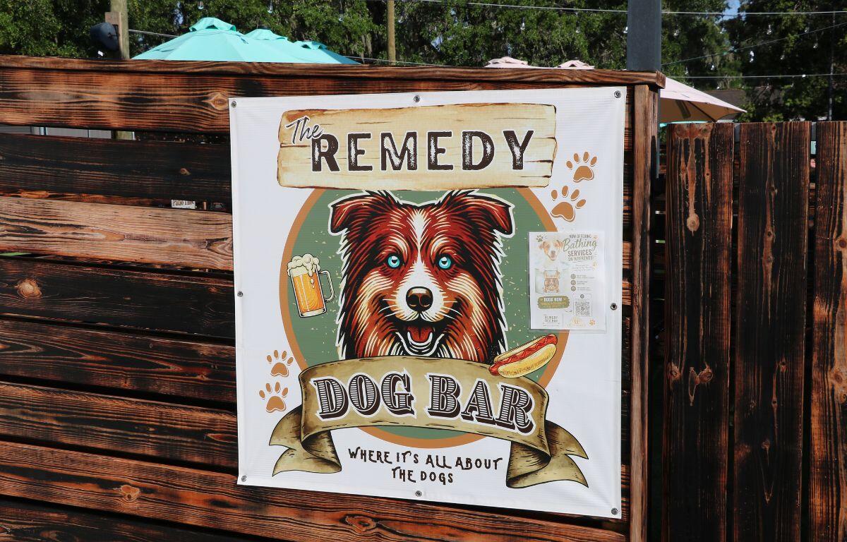 A white sign on a wooden fence with a picture of a dog and text reading, "The Remedy Dog Bar Where It's All About the Dogs."