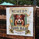 A white sign on a wooden fence with a picture of a dog and text reading, "The Remedy Dog Bar Where It's All About the Dogs."