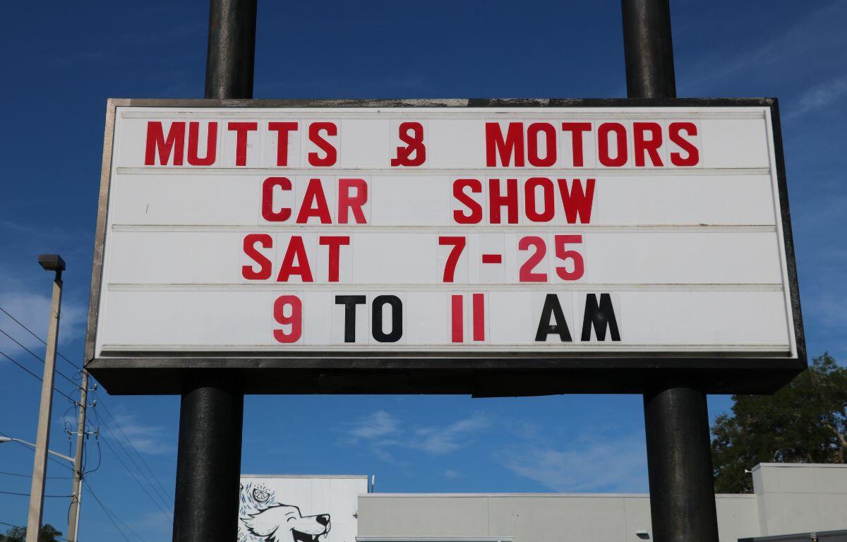 Sign advertising a 'Mutts & Motors' car show: Sat 7-25, 9 to 11 AM, outdoor roadside banner against a blue sky.