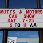 Sign advertising a 'Mutts & Motors' car show: Sat 7-25, 9 to 11 AM, outdoor roadside banner against a blue sky.