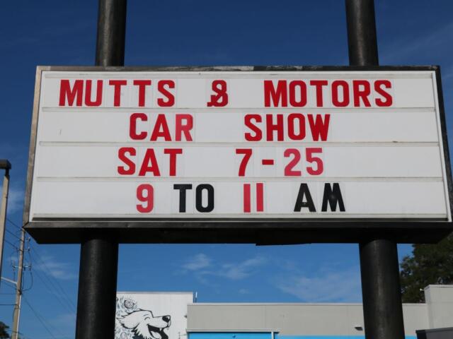 Sign advertising a 'Mutts & Motors' car show: Sat 7-25, 9 to 11 AM, outdoor roadside banner against a blue sky.