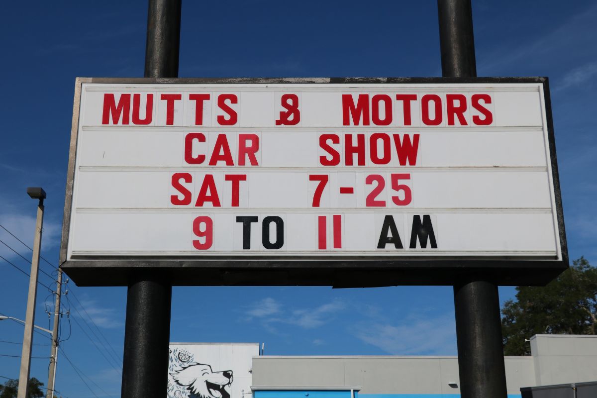 Sign advertising a 'Mutts & Motors' car show: Sat 7-25, 9 to 11 AM, outdoor roadside banner against a blue sky.