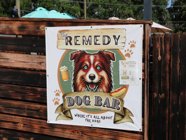 A white sign on a wooden fence with a picture of a dog and text reading, "The Remedy Dog Bar Where It's All About the Dogs."