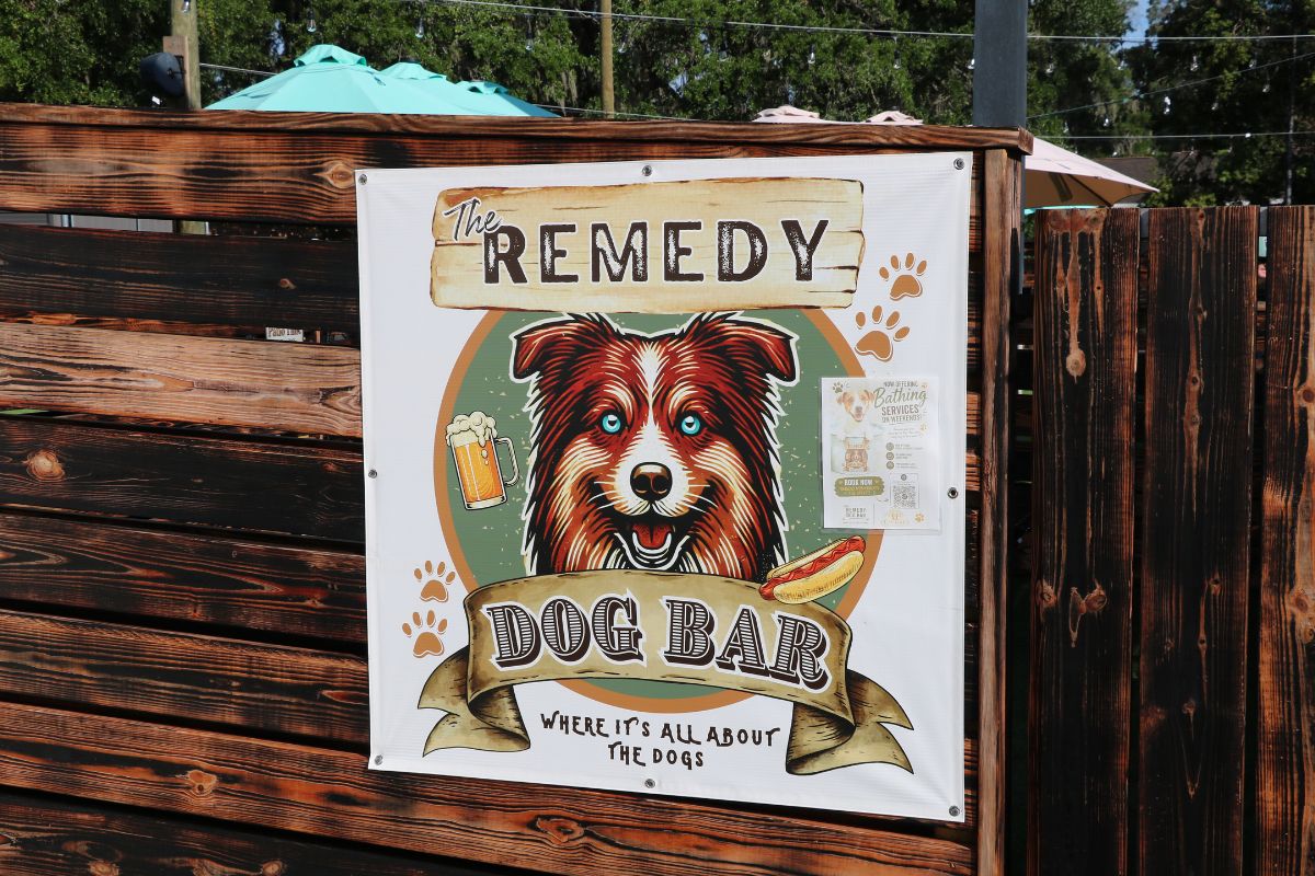 A white sign on a wooden fence with a picture of a dog and text reading, "The Remedy Dog Bar Where It's All About the Dogs."