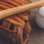 Baseball glove with a wooden bat resting on a wooden surface beside a white baseball.