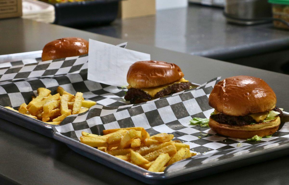 Three burgers with cheese and lettuce on toasted buns, plus French fries on a metal tray lined with black-and-white paper in a diner setup.