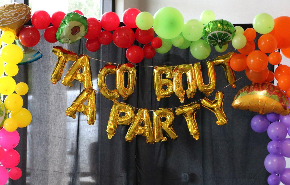 Colorful backdrop with a rainbow balloon arch and gold foil letter banner against a dark curtain, plus a taco-shaped balloon on the side.